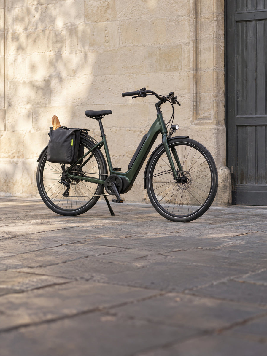 Vélo électrique vert forêt avec sacoche et baguette, garé contre un mur en pierre blonde à Bordeaux